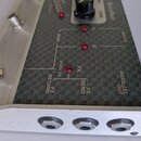 Detailní pohled na ovládací panel efektového pedálu s tmavým vzorovaným povrchem a stříbrným kovovým rámem. Na panelu jsou vidět červené kontrolky s…