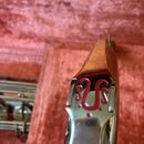 Detailní pohled na kovovou část hudebního nástroje v červeném pouzdře. Na povrchu je umístěno červené dekorativní logo ve tvaru stylizovaného symbolu…