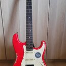 Červená elektrická kytara typu Stratocaster s bílým pickguardem a hnědým hmatníkem. Na hlavě je vidět nápis "Squier". Tělo nástroje vykazuje výrazné…