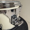 Detailní pohled na bílý buben s chromovaným obručem a kování. Na kovovém držáku je vyryto logo „Gretsch“. Povrch chromu vykazuje drobné oděrky a znám…
