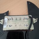 Fotografie zachycuje spodní stranu elektrického snímače s kovovým krytem, který je umístěn na černém plastovém pickguardu. Na spodní straně krytu je…
