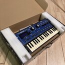 Modrý syntetizátor s klaviaturou umístěný v přepravním kartonu s bílým polystyrenem. Na přístroji je nápis MININOVA a logo novation. Zařízení obsahuj…