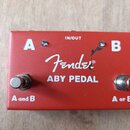Červený kovový efekt pedál s bílými nápisy. Uprostřed je logo Fender® a text ABY PEDAL, v horní části nápis IN/OUT a po stranách písmena A a B. Na sp…