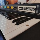 Detailní pohled na klávesový nástroj s černým povrchem a bílými a černými klávesami. Na horním panelu je viditelný nápis PIANO v obdélníkovém rámečku…