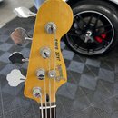 Zadní strana hlavy basové kytary z lehce žlutého dřeva. Na povrchu je černý nápis „Fender® JAZZ BASS“. Jsou vidět čtyři chromované ladičky. Povrch dř…