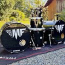 Černá bicí souprava složená z dvou basových bubnů, jednoho tom-tomu a jednoho floor tomu. Na předních blanách basových bubnů je bílý nápis METAL FACT…