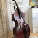 Tmavě hnědá kontrabasová violoncello v lakovaném provedení, umístěná na černém stojanu. Nástroj má dřevěné tělo s tradičním tvarem, čtyři struny a sv…