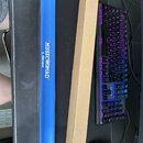Modrá anodizovaná hliníková lišta s bílým nápisem MUSICNOMAD™ L-Beam. Vedle ní leží úzký hnědý kartonový obal s pomačkaným koncem. V pozadí je vidět…