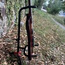 Profilní pohled na hnědý smyčcový nástroj umístěný v černém stojanu s červeným základním křížem. Nástroj má lakovaný dřevěný povrch s viditnou strukt…