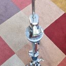 Stříbrný kovový pedál na hi-hat značky MAPEX. Nástroj stojí na vícebarevném koberci a je vybaven kovovou tyčí a mechanismem s utahovacími šrouby. Pov…