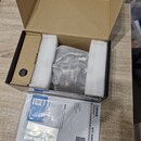 Otevřená kartonová krabice s bílým polystyrenem, ve které je v průhledné fólii zabalený černý přístroj. Před krabicí leží papírová dokumentace s nápi…