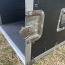Detail černého transportního kufru s hliníkovými hranami. Na boku je vidět kovový zámek a úchytka, které vykazují známky oxidace a povrchové koroze.…