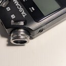 Černý digitální záznamový zařízení s displejem. Na těle jsou viditelné nápisy TASCAM, REC, PEAK a Ω/LINE OUT, v horní části je částečně vidět označen…
