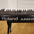 Černý digitální klavír umístěný na černém stojanu. Na přední straně je bílý nápis Roland JUNO-D. Horní panel obsahuje tři posuvníky a několik ovládac…