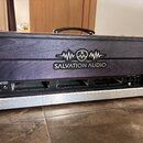Černý transportní kufřík s dřevěným předním panelem a hliníkovým rámem. Na panelu je bílé logo s nápisem SALVATION AUDIO a grafickým symbolem vlny. V…
