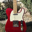 Červená elektrická kytara typu Telecaster s bílým pickguardem a světle hnědým hmatníkem z javoru. Nástroj je umístěn na černém stojanu v venkovním pr…