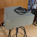 Šedý kovový box s hranatým tvarem, na kterém je položen černý kabel. Na boku boxu jsou vidět čtyři stříbrné šrouby a v levé části vyčnívá kulatý ovlá…