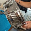 Fotografie zachycuje chromovaný malý buben, který drží osoba v rukou. Nástroj má lesklý kovový povrch a viditelný mechanismus napínání. Na těle nástr…