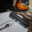 Černý efekt pedál s žebrovaným horním povrchem a bílým štítkem. Do přístroje je zapojen bílý kabel s nápisem Fender a nečitelným textem v jiném písmu…