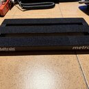 Černý kovový pedalboard s povrchem z černé textury. Na přední hraně je vlevo bílý nápis pedaltrain® a pod ním drobným písmem U.S. Pat. No. 6,458,023…