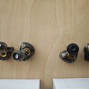 Fotografie zobrazuje čtyři In-Ear monitory s průhledným tmavě šedým plastovým tělem a zlatými vnitřními komponenty. Dva sluchátka mají nasazené světl…