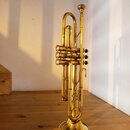 Zlatavý mosazný trumpet s třemi ventily. Nástroj stojí vertikálně na dřevěném stole. Na povrchu nejsou vidět žádné značky, loga ani nápisy. Povrch vy…