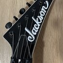 Fotografie zachycuje hlavu elektrické kytary v černé barvě s bílým lemováním. Na povrchu je umístěno bílé logo „Jackson“. Nástroj má šest černých lad…