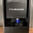 Zadní strana černého kytarového komba s texturovaným povrchem. Na zadním panelu je bílé logo tc electronic a v horní části varovný štítek s texty v n…