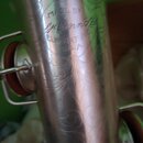 Detail stříbrného kovového nástroje s rytým ornamentem. Na povrchu je vyryt nápis MADE BY C.G. Conn Ltd ELKHART IND. U.S.A. Povrch vykazuje drobné šk…