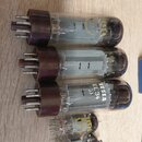 Fotografie zobrazuje skupinu elektronických vakuových lamp s hnědými plastovými základnami, položených na dřevěném povrchu. Tři největší lampy mají n…