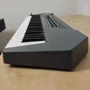 Šedý elektronický klavír s černobílými klávesami. Na horním ovládacím panelu jsou vidět posuvníky a tlačítka. Povrch nástroje je matný, bez viditelný…