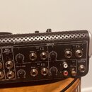 Záběr na zadní panel černého audio zařízení s kovovým perforovaným krytem v horní části. Na panelu jsou vidět různé konektory, potenciometry a nápisy…