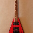 Červená elektrická kytara s ostrým, V-tvarovým korpusem a černým lemováním. Na hlavě hmatníku je bílý nápis Jackson. Nástroj má dva humbuckery, jeden…