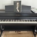 Černý digitální piano s klávesnicí a třemi pedály. Na přední straně korpusu je zlatý nápis KAWAI. Na notovém pultu je umístěn časopis s portrétem a t…