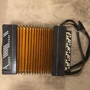 Černý akordeon s oranžovými měchy a bílými knoflíky. Na pravém černém krytu je svisle napsán nápis Hohner. Nástroj má černé popruhy a je položen na b…