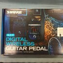 Fotografie znázorňuje krabici s produktem Shure GLX-D Digital Wireless Guitar Pedal. Na obalu je zobrazen černý bezdrátový pedál s červeným digitální…