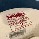 Detailní záběr na povrch kovového činele zlatavé barvy. Na povrchu je červený nápis v rámečku: „PAiste 2002“, pod ním text „TRADE MARK“ s hvězdou v k…