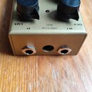 Zlatý efekt pedál s čtyřmi černými oválnými knoflíky a stomečem. Na horní straně jsou nápisy GAIN, IN, MADE IN USA, 9VDC a OUT. Na spodní hraně jsou…