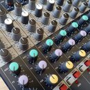 Detailní pohled na ovládací panel míchacího pultu s řadami šedých otočných potenciometrů. Na panelu jsou bílé nápisy AUX1, FX a PAN. Povrch pultu je…