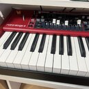 Červený digitální klavír s bílými a černými klávesami. Na horním panelu je bílý nápis nord stage 4 a pod ním menší text NORD KEYBOARD. Přístroj obsah…