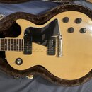 Světle žlutá elektrická kytara s černým pickguardem a čtyřmi ovládacími knoflíky v hnědém plišovém pouzdře. Nástroj má dva černávé humbuckery a stříb…
