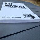 Bílá kniha s černým textem a grafikou, položená na šedém povrchu. Na obálce je nápis KLF MANUAL a pod ním v závorce (JAK SE DOSTAT NA VRCHOL HITPARÁR…