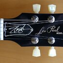 Fotografie zachycuje hlavu černé elektrické kytary s bílými ladicími kolíky. Na povrchu je bílý nápis Gibson, podpis Les Paul a slovo MODEL. Vlevo je…
