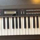 Černý klávesový syntetizátor s bílými a černými klávesami. Na horním panelu jsou nápisy Roland a JUNO-1, dále text PROGRAMMABLE POLYPHONIC SYNTHESIZE…
