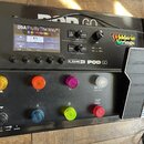 Černý kytarový procesor s displejem a expresním pedálem. Na přístroji je bílý nápis LINE 6 POD GO a nálepka D'Addario Strings. Displej zobrazuje text…