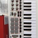 Bílá MIDI klaviatura s šedými klávesami a ovládacími prvky. Na přístroji je nápis MPK mini a logo AKAI PROFESSIONAL. Obsahuje 8 šedých padů, osmtočac…