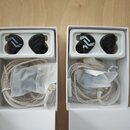 Dvě bílé krabičky s černými in-ear monitory. Sluchátka mají v plastovém lůžku stříbřité kabelové vedení a jsou připojena k bílým opleteným kabelům. N…