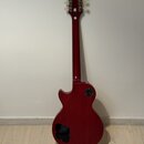 Zadní pohled na červenou elektrickou kytaru s tvarem typu Les Paul. Na zadní straně hlavy je vidět malý bílý štítek s nečitelným textem. V dolní část…