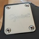 Detailní záběr na kovový, chromovaný kryt zadní části elektrokitary s nápisem Squier®. Kryt je připevněn čtyřmi šrouby k černému lakovanému povrchu n…