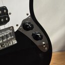 Detail elektrické kytary s černým lakovaným korpusem a bílým pickguardem. Na černé plastové části jsou umístěny dva černé oválné ovládací knofíky. V…